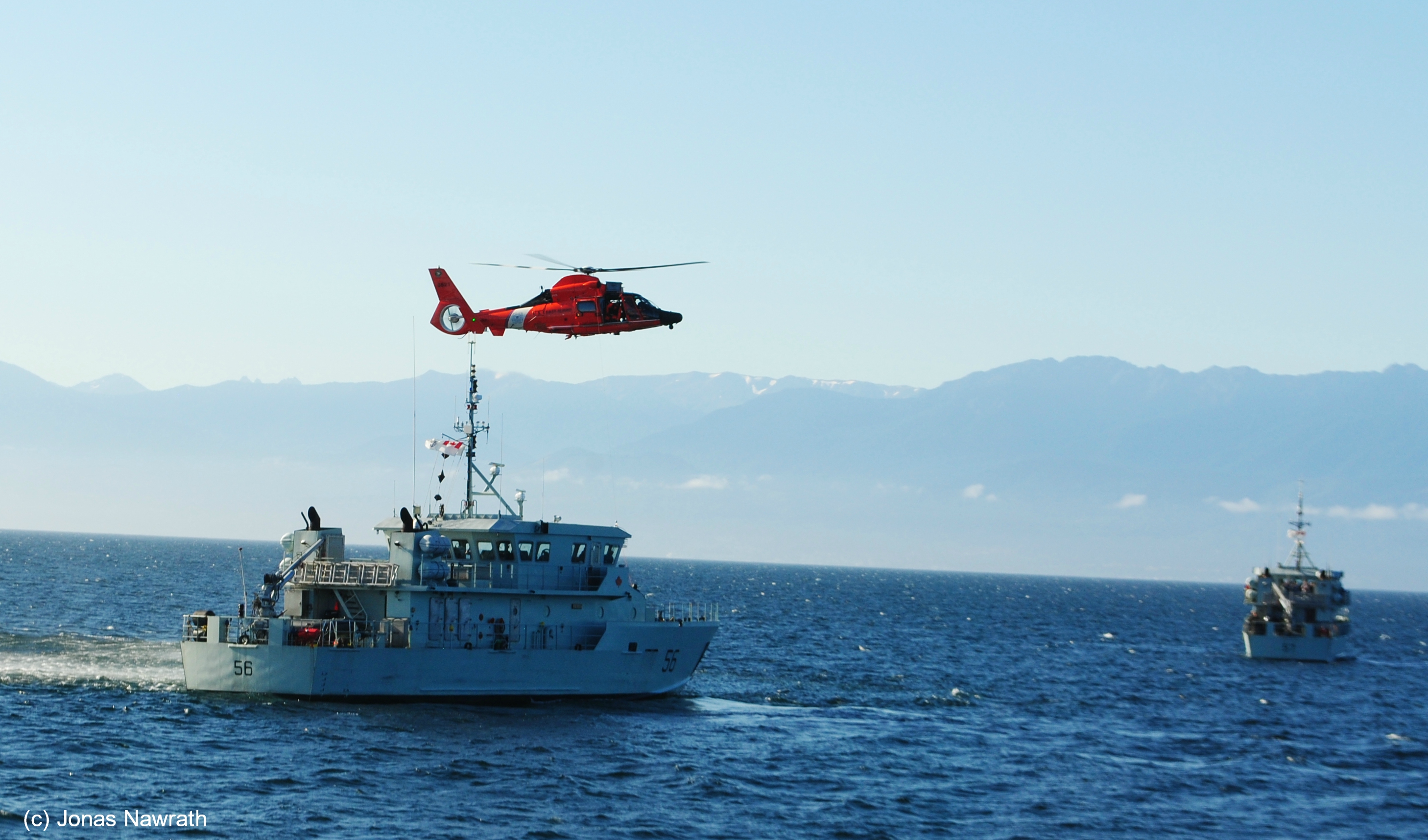 A red military helicopter flies over a military vessel